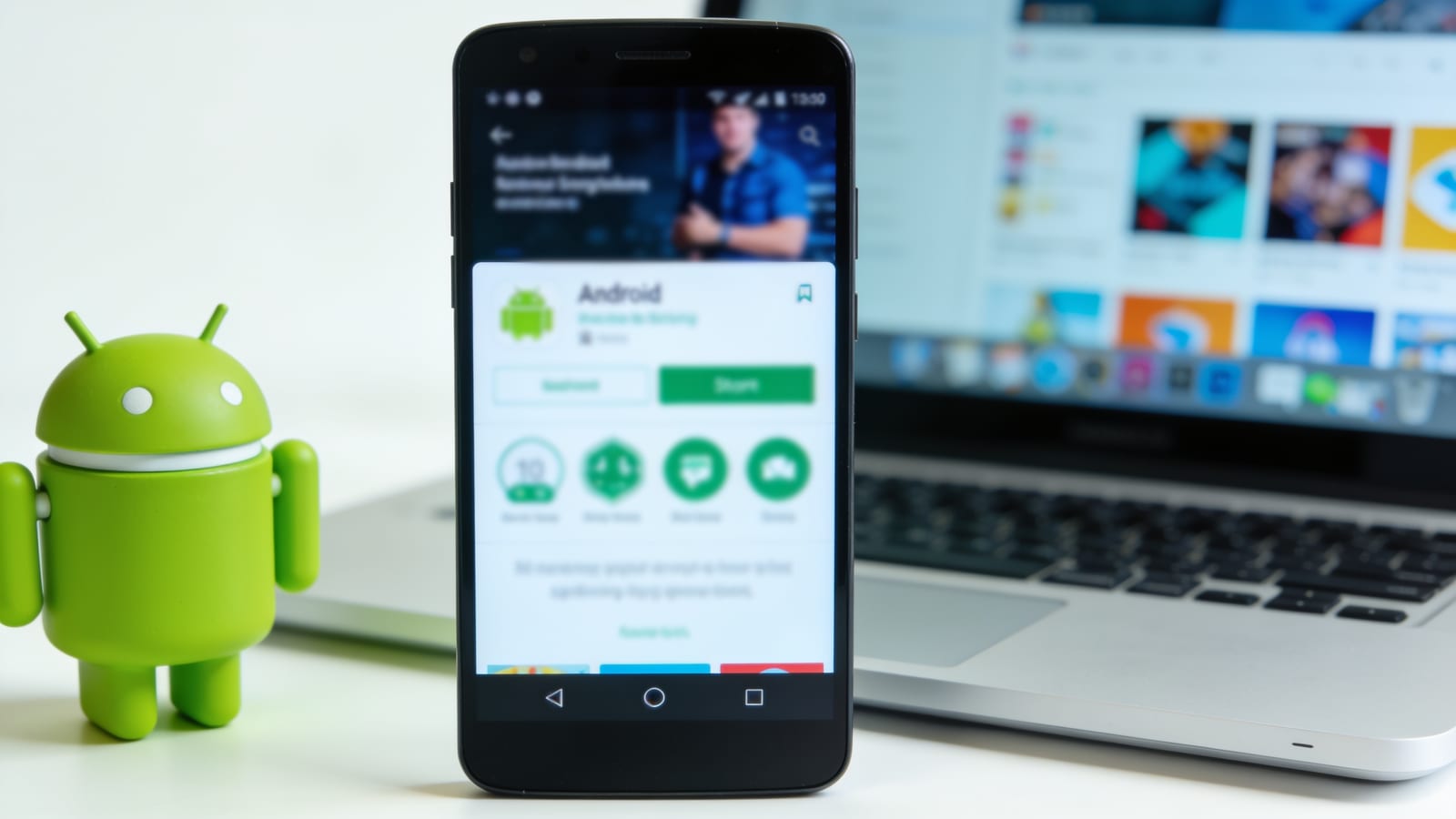 Smartphone Android posé sur un bureau clair à côté d'une figurine du robot Android vert et d'un ordinateur portable, esthétique épurée